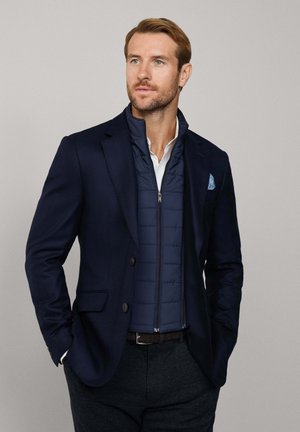 Blazer blu navy con due bottoni, indossato sopra un gilet imbottito blu navy con chiusura a zip. Camicia bianca sotto; pantaloni grigi strutturati.