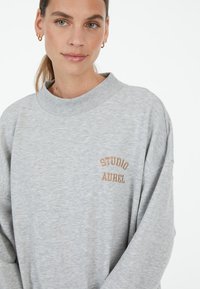 Gray sweatshirt with a rounded neckline and long sleeves. Features an embroidered "STUDIO AUREL" in brown on the front. Soft texture.