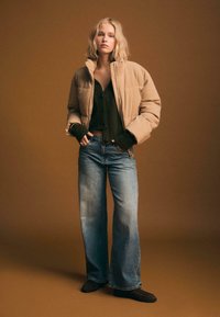 Beżowa kurtka puchowa z wysokim kołnierzem, noszona na ciemnym swetrze, zestawiona z jasnoniebieskimi spodniami typu wide-leg oraz ciemnymi botkami za kostkę.