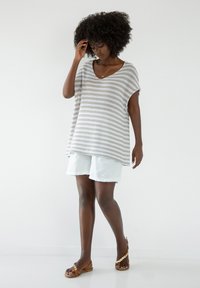 Skrzyżowany sweter w jasnym beżu i bieli, w połączeniu z jasnymi szortami. Top ma luźny krój i krótkie rękawy. Noszony z brązowymi sandałami.