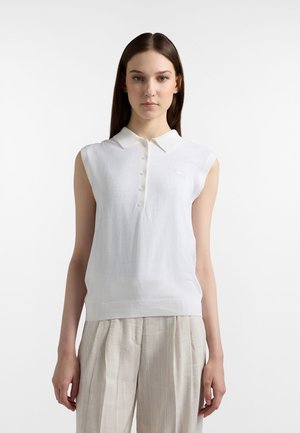 Młoda kobieta o prostych, brązowych włosach, ubrana w bezrękawnik w białym kolorze z kołnierzykiem i jasnobeżowe plisowane spodnie.