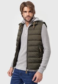 Olivengrøn puffervest med lynlås foran, to sidelommer med lynlås og hætte. Båret over en lysegrå strikket sweater, parret med blå jeans.