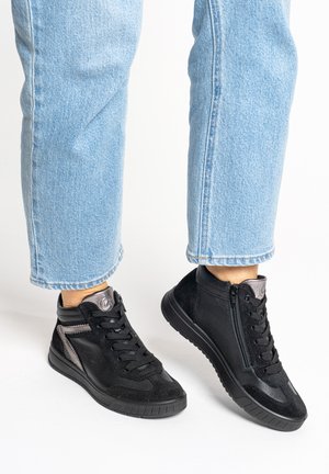 Personne portant un jean bleu clair et des baskets montantes noires avec des fermetures éclair latérales sur fond blanc.