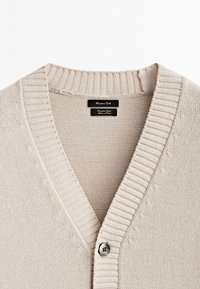 Cardigan beige tricoté avec col en V côtelé et un seul bouton gris, montrant les étiquettes de marque et de matière à l'intérieur du col.