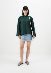 Grønn langermet skjorte med knapper, lys denim miniskjørt med frynset kant, og svarte sandaler med teksturert mønster, mot en ensfarget bakgrunn.