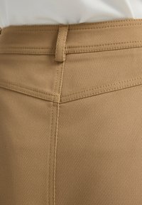Bruine, gestructureerde stof met een diagonaal patroon; zichtbare stiksels en een riemlus bij de taille. Gerichte weergave van het bovenste gedeelte van het kledingstuk.