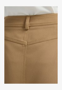 Tela marrón texturizada con un patrón diagonal; costuras visibles y un lazo para el cinturón en la cintura. Vista enfocada en la sección superior de la prenda.