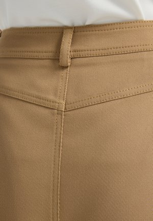 Ruskea, teksturoitu kangas, jossa on viistokuvio; näkyvissä ompelujälki ja vyölenkki vyötäröllä. Tarkennettu näkymä vaatteen yläsosasta.