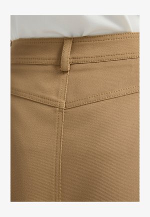 Ruskea, teksturoitu kangas, jossa on viistokuvio; näkyvissä ompelujälki ja vyölenkki vyötäröllä. Tarkennettu näkymä vaatteen yläsosasta.