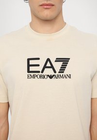Béžové bavlněné tričko s krátkým rukávem, na němž je uprostřed hrudi výrazné černé logo "EA7 EMPORIO ARMANI".