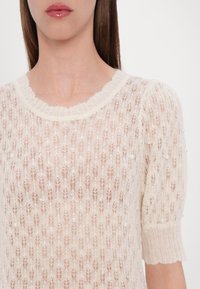 Gros plan d'une personne portant un haut tricoté beige clair avec des manches courtes bouffantes et de petites décorations en perles.