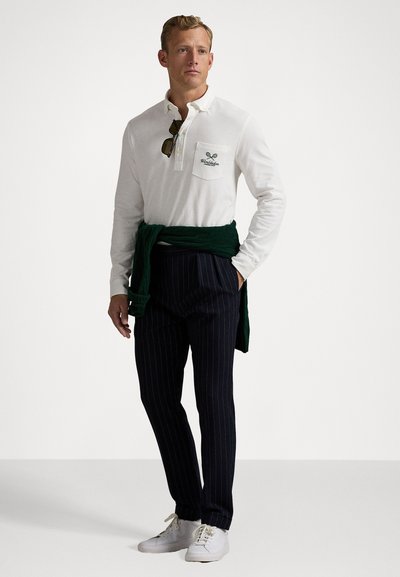 Polo alb cu mâneci lungi și logo pe buzunar, asortat cu pantaloni în dungi închise. Pulover verde legat în jurul taliei, adidași albi.