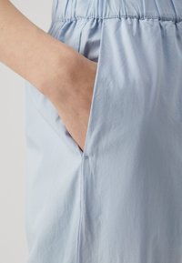 Lichtblauwe stof met een zachte textuur, voorzien van een zijzak en een gerimpelde tailleband. Het materiaal lijkt licht en glad te zijn.