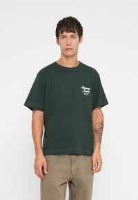 Dark green cotton t-shirt with short sleeves, crew neck, and a white logo on the left chest. Smooth texture with a relaxed fit.