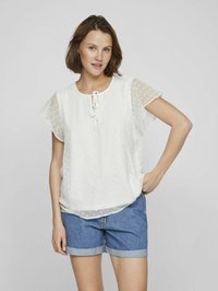 Blusa bianca a maniche corte con pois, colletto rotondo e dettaglio con fiocco. Abbinata a pantaloni shorts di jeans con risvolti.