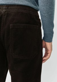Hand resting near the side of dark brown corduroy pants with a back pocket, worn with a blue long-sleeve sweater.