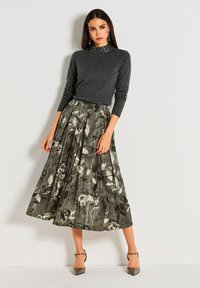 Frau mit langen dunklen Haaren, die ein dunkelgraues Langarmoberteil und einen gefältelten, floral gemusterten Midirock trägt, steht vor einer weißen Wand.