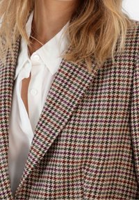 Texturerad kavaj i houndstooth-mönster med nyanser av grått, vinrött och lila. Bärs över en vit skjorta med krage.