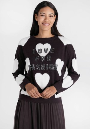 Vrouw draagt zwarte trui met witte hartjes en pailletten tekst "LOVE FOR FASHION,", gecombineerd met een zwarte geplooide rok, glimlachend met gevouwen handen.