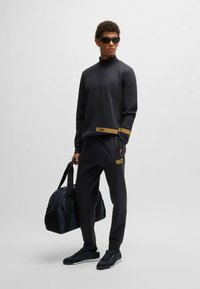 Mörkgrå sportig sweatshirt med hög krage, guldaccenter vid fållen. Matchas med byxor i samma färg och en marinblå duffelväska.