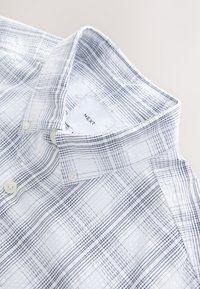 Camicia bianca con chiusura a bottoni, caratterizzata da un motivo a quadri blu e grigio. Tessuto di cotone morbido con colletto classico e due bottoni di chiusura.