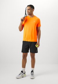 Bright orange short-sleeve sports shirt, black shorts, holding a green tennis ball, with a tennis racket resting on his shoulder. White sneakers.