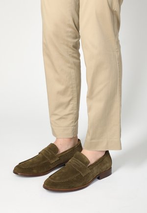 Samt-Loafer in Olivgrün mit einer gesteppten Akzentnaht und einem niedrigen Holzabsatz, kombiniert mit beige schmal geschnittenen Hosen.