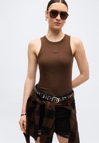 Νεαρή γυναίκα με καφέ τοπ χωρίς μανίκια, μαύρη ζώνη με καρφιά, πουκάμισο καρό δεμένο στη μέση, μαύρο σορτς και μεγάλα σκοτεινά γυαλιά ηλίου.