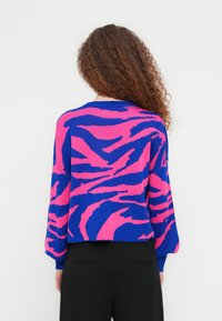 Γυναίκα με σγουρά μαλλιά, ντυμένη με πουλόβερ σε μπλε και ροζ αφηρημένο σχέδιο και μαύρο παντελόνι, στέκεται αντικρίζοντας μια λευκή τοιχοποιία.