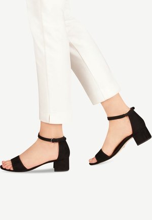 Czarne zamszowe sandały na słupku z paskiem na kostce, otwartym noskiem i gładką fakturą. Noszone z białymi, krótkimi spodniami.