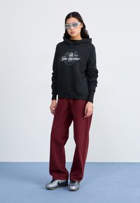 Hoodie preto com logotipo branco, calças largas cor bordeaux e sapatos metálicos prateados. O modelo usa óculos de sol cinzentos. Fundo simples.