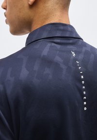 Άνδρας που φοράει σκούρο μπλε πουκάμισο με γιακά, με διακριτικά σχέδια και κάθετο λευκό κείμενο "J.LINDEBERG" στην επάνω πλάτη.