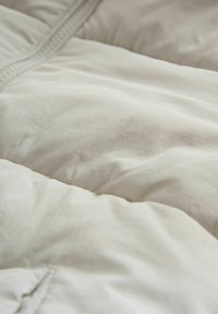 Svijetla puf jakna s kvadratnim uzorkom, glatkom teksturom i suptilnim sjajem; ima vidljive šavove i detalj sa zatvaračem.