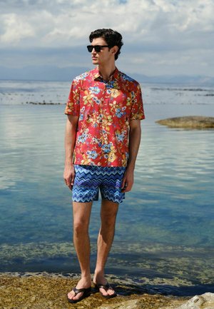 Mann mit Sonnenbrille, rotem Blumenhemd, blauen gemusterten Shorts und Flip-Flops steht an einem felsigen Ufer am ruhigen Meer unter bewölktem Himmel.