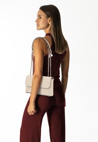 Bolso de mano de cuero beige con forma estructurada, que presenta cadenas plateadas y un cierre de solapa. Textura suave con bordes negros contrastantes.