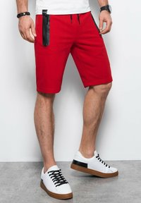 Shorts rouges arrivant au genou avec des zips noirs sur les côtés, fabriqués à partir d'un tissu lisse. Associés à des baskets blanches avec des accents noirs et des semelles brunes.