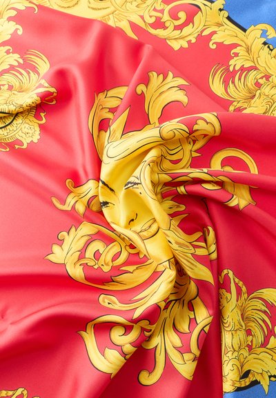 Κόκκινο ύφασμα με περίτεχνα μοτίβα μπαρόκ σε κίτρινο-χρυσαφί και σχεδιασμό με ηλιακό πρόσωπο σε στυλιζαρισμένη μορφή, απεικονίζεται με φυσικές πτυχώσεις και υφή.