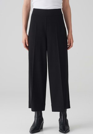 Zwart wijde pantalon met een gladde textuur, voorzien van diepe plooien, een hoge tailleband en een nette afwerking aan de zoom. Gepaard met zwarte enkellaarzen.