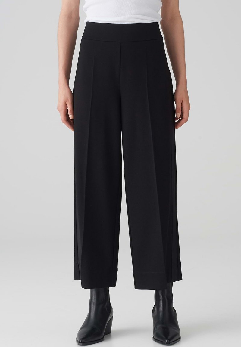 Pantalons larges noirs avec une texture lisse, présentant de profonds plis, une taille haute et une finition nette à l'ourlet. Associés à des bottines noires.