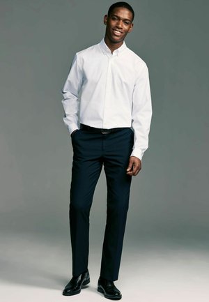 Chemise blanche à boutons associée à un pantalon foncé. La chemise présente un col classique ; le pantalon est cintré avec une texture lisse. Des chaussures noires complètent le look.