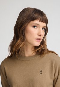 Beiger Pullover mit Rundhalsausschnitt und gerippten Bündchen, mit kleinem gesticktem Logo auf der Brust. Weiche Strickstruktur.