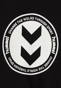 Svart stoff med et sirkulært hvitt logo som har to nedoverpekkende vinkelstreker og teksten "hummel" og "ENDRE VERDEN GJENNOM SPORT."