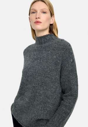 Kobieta o jasnych włosach, ubrana w ciemnoszary, miękki sweter z guzikami wzdłuż lewego ramienia, stojąca na jednolitym tle.