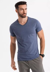 Šedé tričko s výstřihem do V vyrobené z bavlny, má krátké rukávy a hladký povrch. Nosit s šedými šortkami a černými hodinkami.