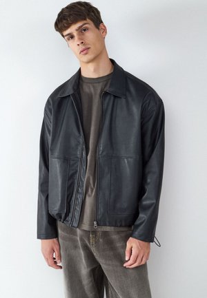 Giovane uomo che indossa una giacca di pelle nera con zip sopra una t-shirt grigio scuro e pantaloni marroni larghi davanti a una parete chiara e semplice.