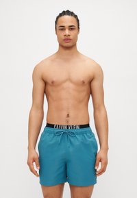 Τεיאור μπάνιο σε τυρκουάζ απόχρωση με κορδόνι και μαύρη ζώνη που φέρει το λογότυπο "CALVIN KLEIN". Ο ιστός φαίνεται λείος και ελαφρύς.