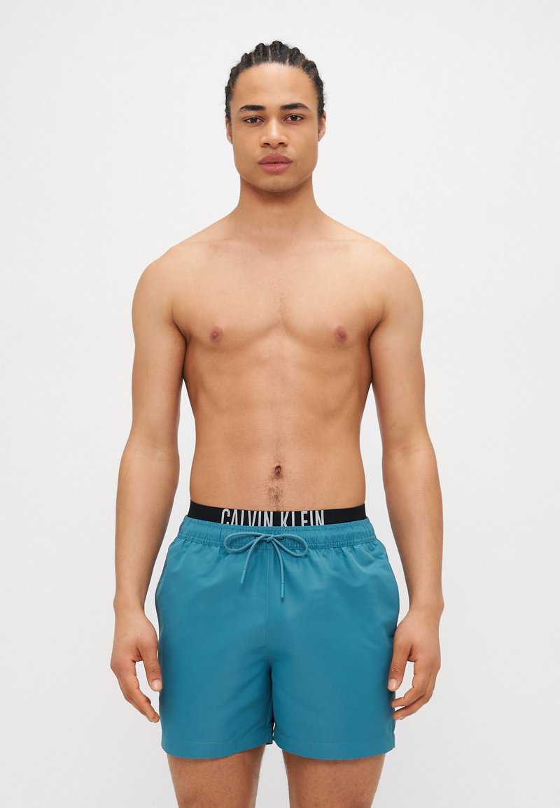 Τεיאור μπάνιο σε τυρκουάζ απόχρωση με κορδόνι και μαύρη ζώνη που φέρει το λογότυπο "CALVIN KLEIN". Ο ιστός φαίνεται λείος και ελαφρύς.