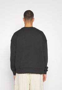 Homme portant un sweat-shirt noir ample et un short cargo beige, vu de dos sur un fond blanc uni.