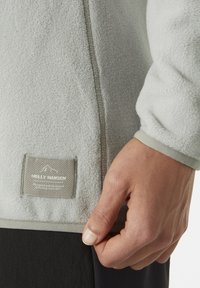 Jasnoszara bluza polarowa o teksturowanej powierzchni, z ciemnym wykończeniem u dołu i metką Helly Hansen blisko dolnej krawędzi.