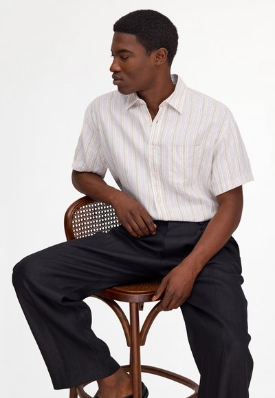 Man sitting on a wooden chair, wearing a short-sleeve light striped shirt and loose black pants, looking to his left.